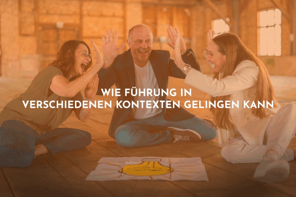 Zwei Frauen und ein Mann sitzen auf dem Boden und strecken die Hände freudestrahlend in die Höhe. Ein Bild einer Glühbirne liegt vor ihnen auf den Boden.