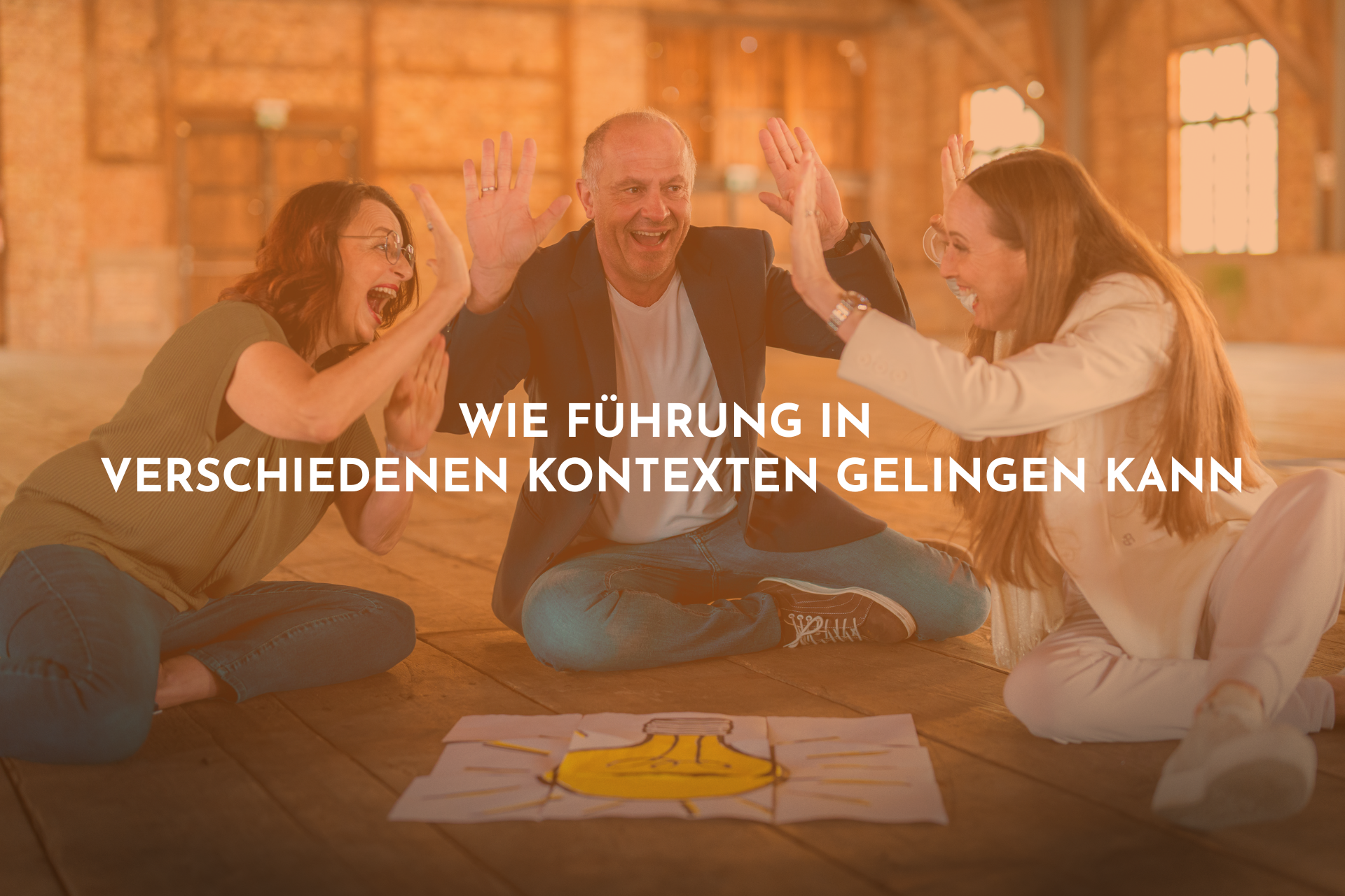 Zwei Frauen und ein Mann sitzen auf dem Boden und strecken die Hände freudestrahlend in die Höhe. Ein Bild einer Glühbirne liegt vor ihnen auf den Boden.