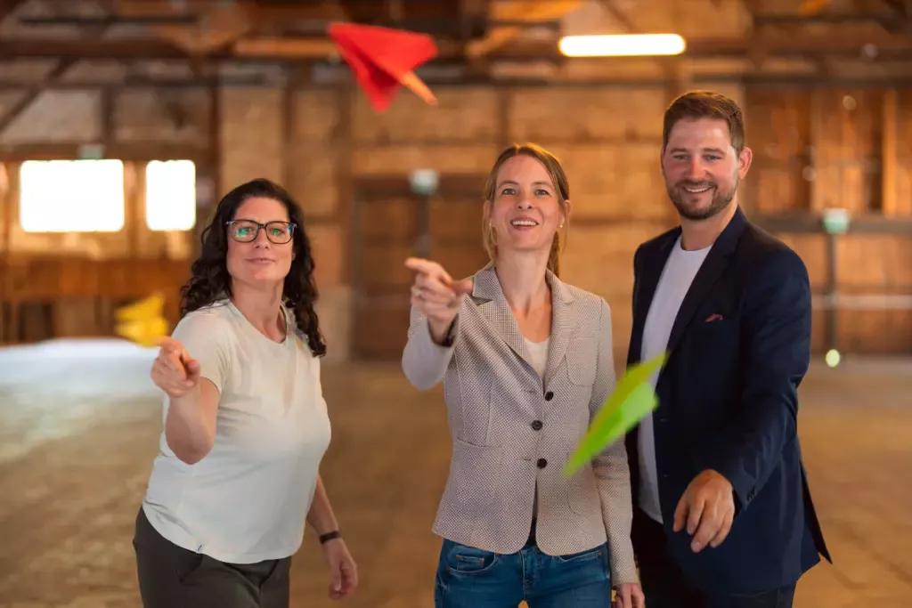 Zwei Frauen und ein Mann lassen bunte Papierflieger fliegen.