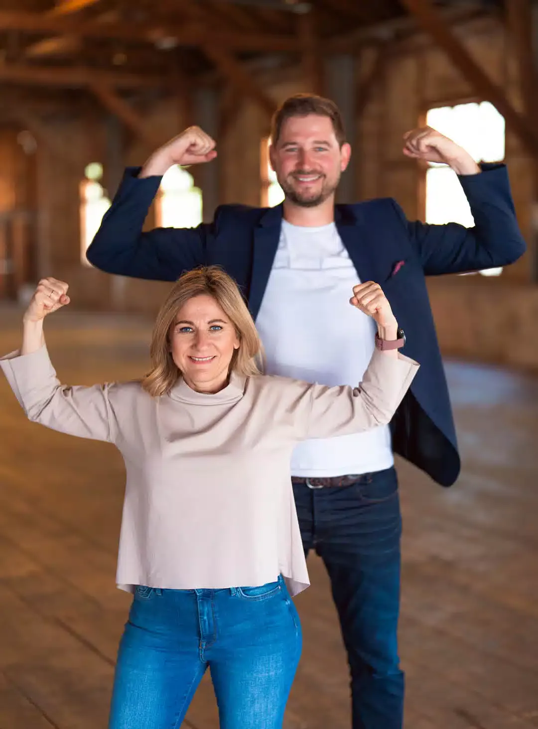 Ein Mann in dunkelblauem Sakko, weißem Shirt und Jeans und davor eine Frau im beigen Langarmshirt und Jeans. Beide heben die Arme und demonstrieren Stärke.