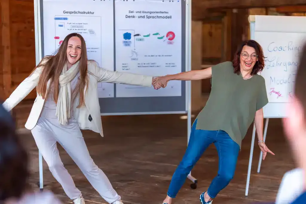 Zwei Damen vor Flipcharts lachen und halten sich an den Händen in der Mitte um Stärke zu zeigen