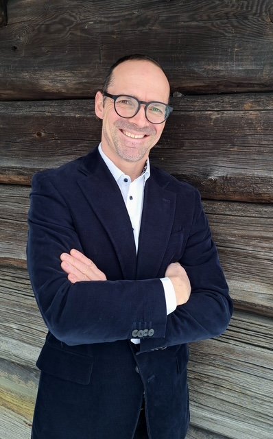 Ein Mann mit Brille im Business Outfit sitzt vor einer Holzwand und schaut freundlich in die Kamera