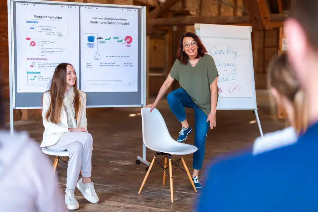 zwei Personen vor einem Flipchart eine Person sitzend und die andere steht und hebt einen Fuß auf den vor ihr stehenden Sessel