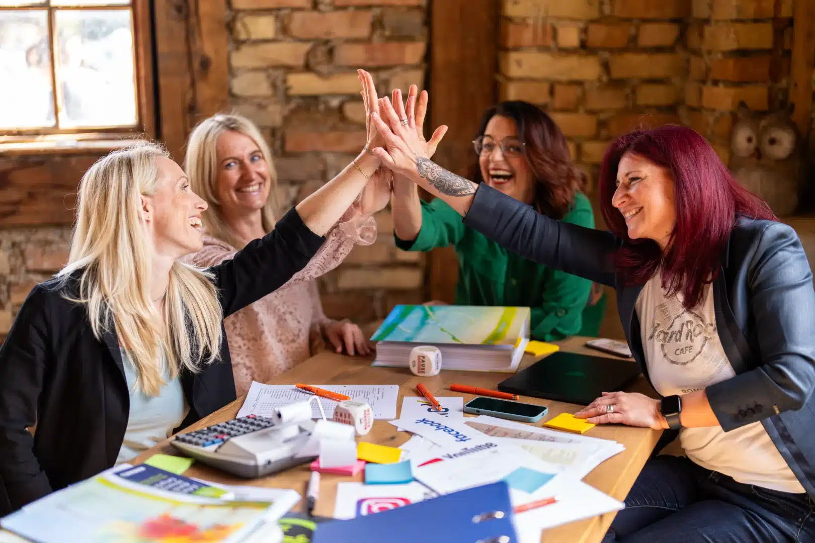 Vier Personen, die an einem Tisch mit Arbeitsmaterialien sitzen. Sie lachen und schlagen in der Mitte die Hände zusammen