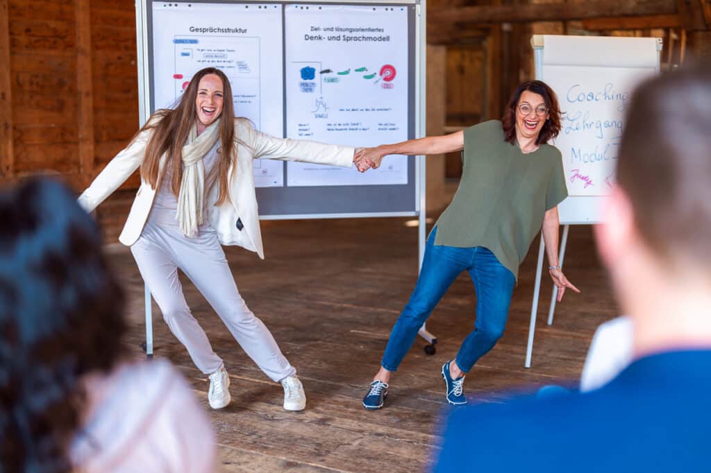 Zwei Damen vor Flipcharts lachen und halten sich an den Händen in der Mitte um Stärke zu zeigen