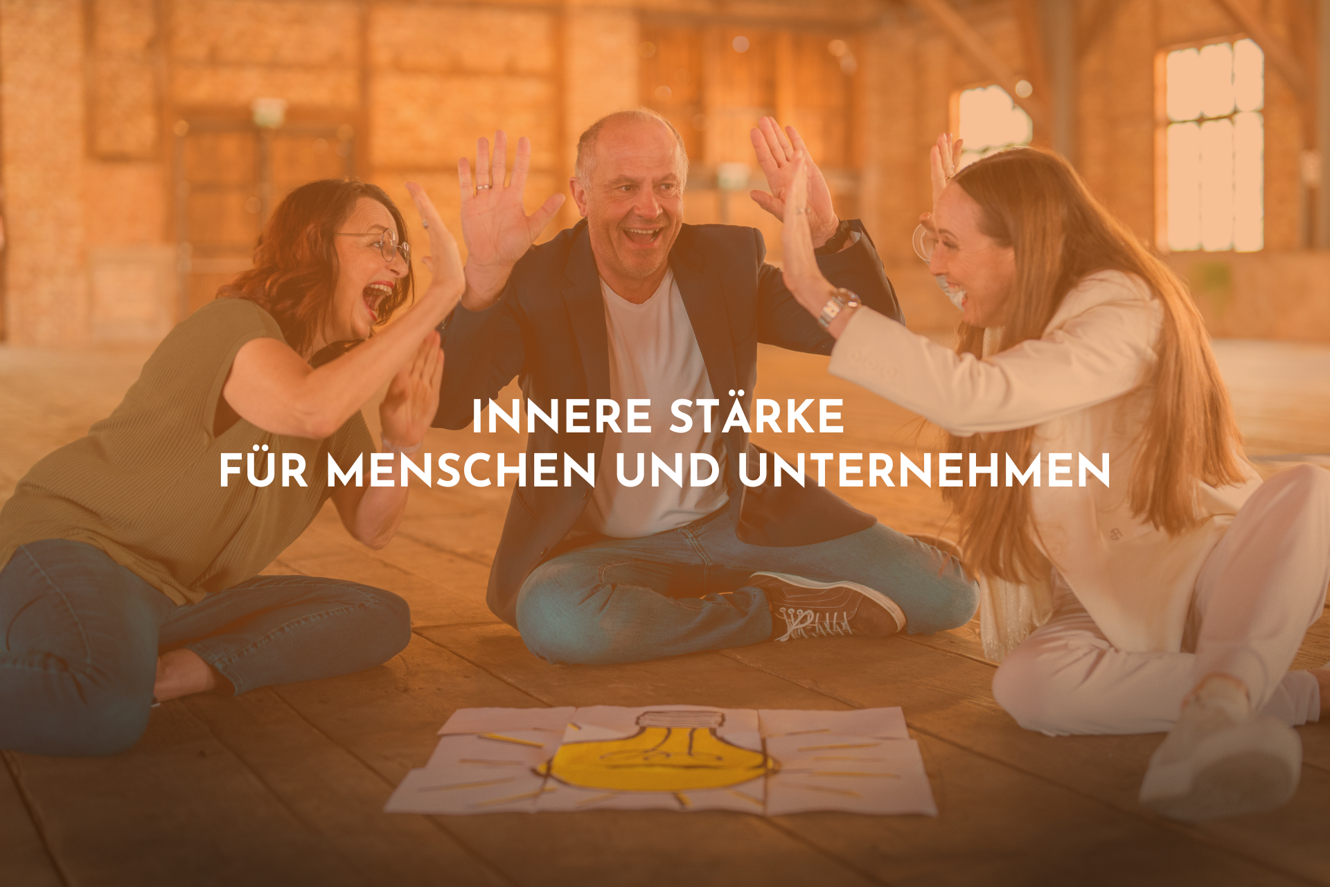 Ein Mann und zwei Frauen sitzen auf dem Boden und freuen sich. Vor ihnen liegt ein großes weißes Blatt Papier mit einer gezeichneten Glühbirne.