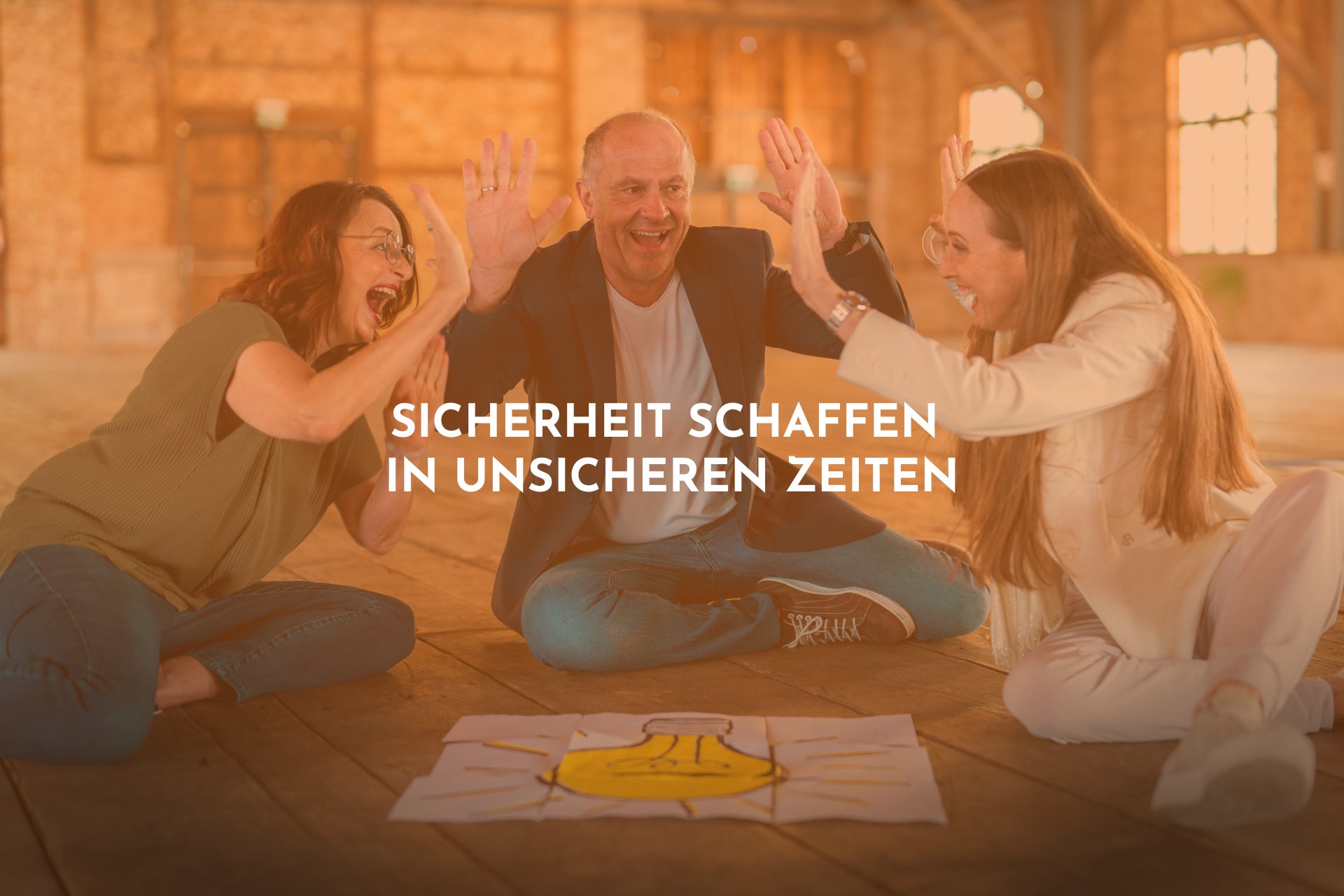 Ein Mann und zwei Frauen schlagen auf dem Boden sitzend und freudestrahlend in die Hände der anderen ein. Vor ihnen liegt ein Bild einer gezeichneten, gelben Glühbirne.